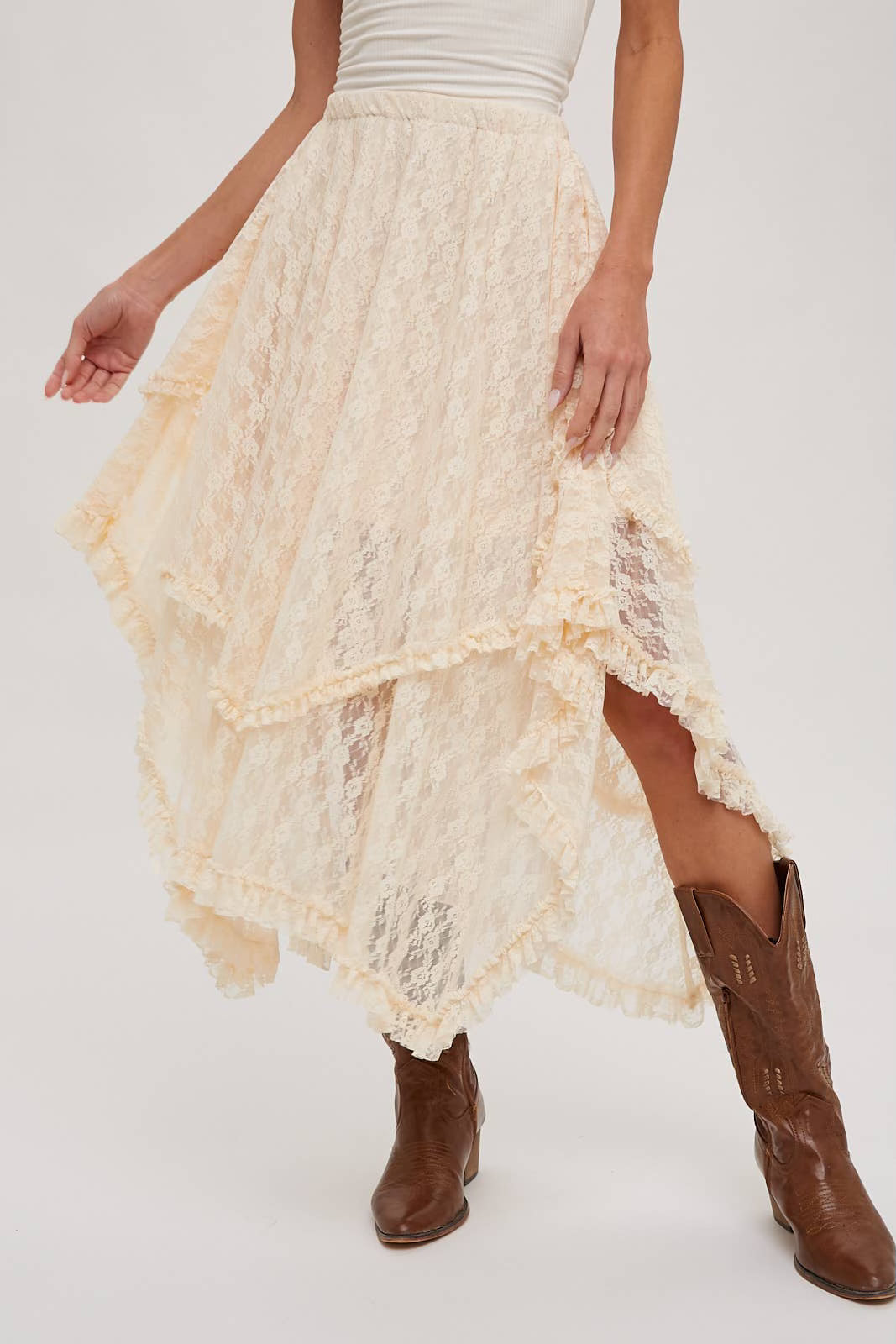 A cream lace high-low skirt with tiered ruffles, styled with brown cowboy boots against a plain white studio background.