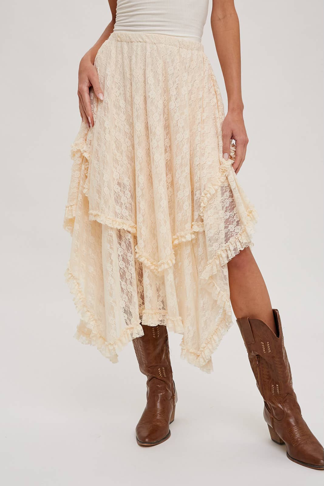 A model wearing a cream lace high-low midi skirt with ruffled layers and brown leather cowboy boots, shown from the waist down against a light gray studio background.