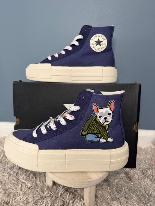 A pair of custom high-top sneakers in navy blue canvas, styled on a small wooden stool in front of a black shoebox. The front shoe features an embroidered French Bulldog wearing a green sweater, while the back shoe displays the Converse-style circular star patch.