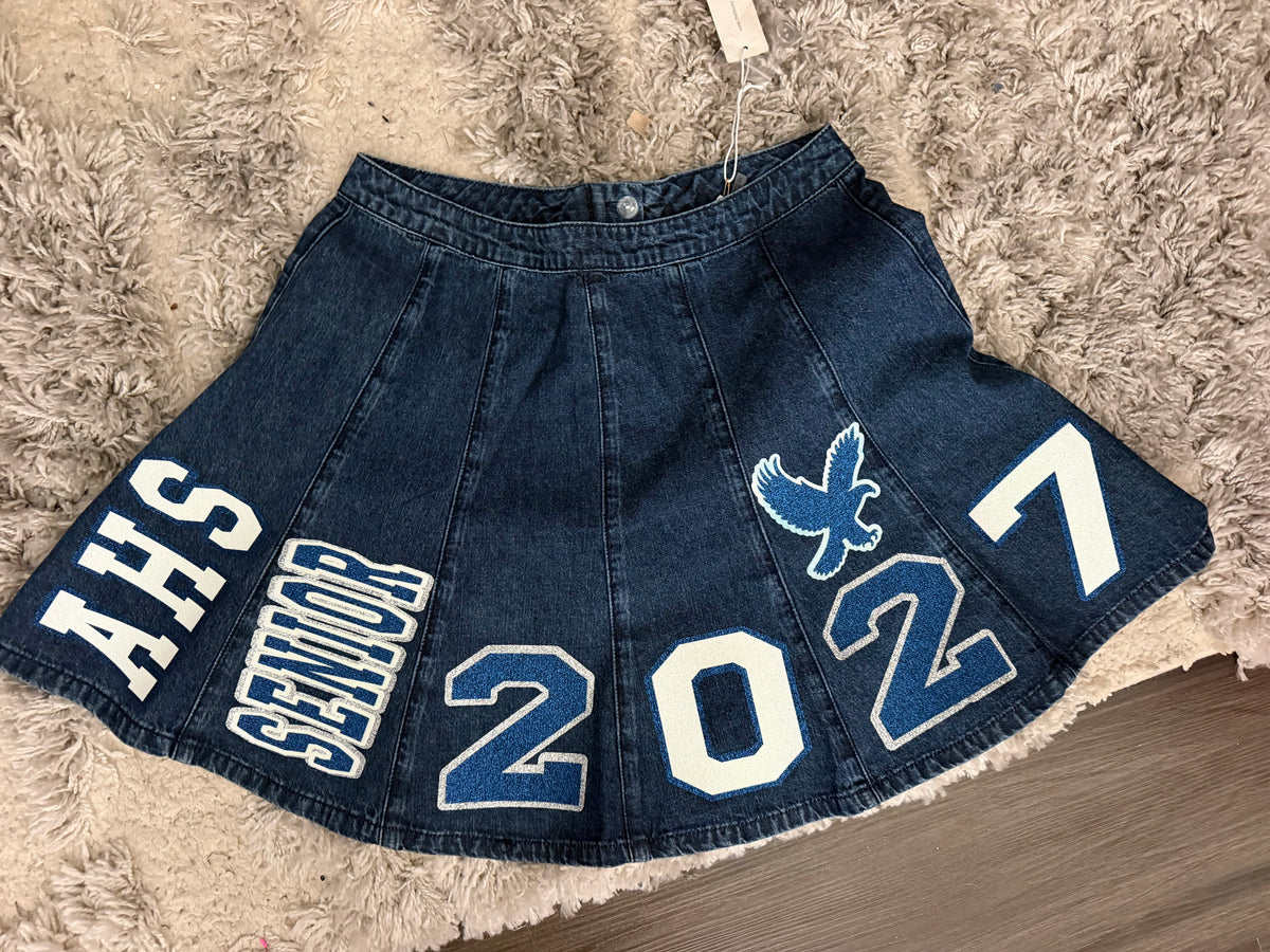 A dark blue denim pleated skirt with white and blue varsity-style lettering and numbers reading 'AHS SENIOR 2027' and a falcon ogo, laid flat on a shaggy beige rug.