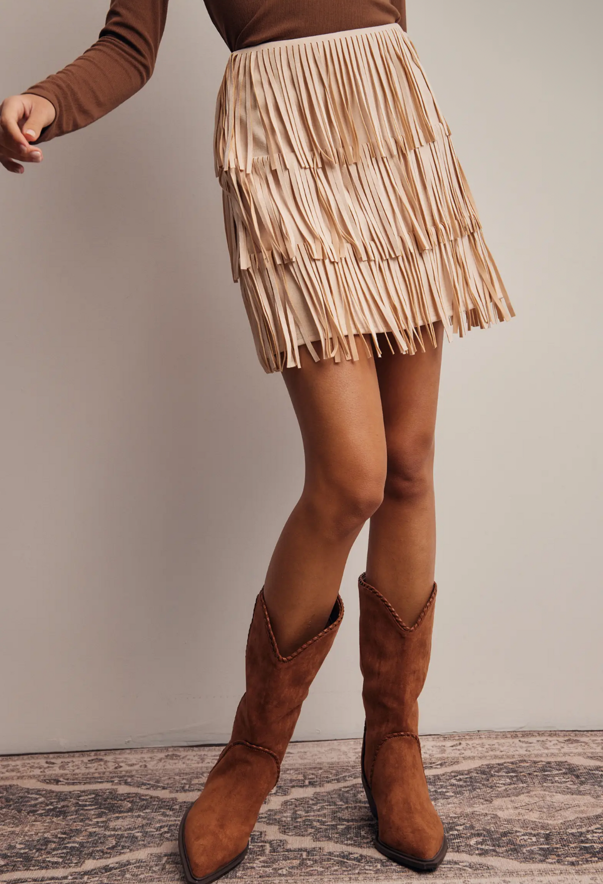 woman wearing a brown top and champagne fringe skirt with brown boots on a neutral background