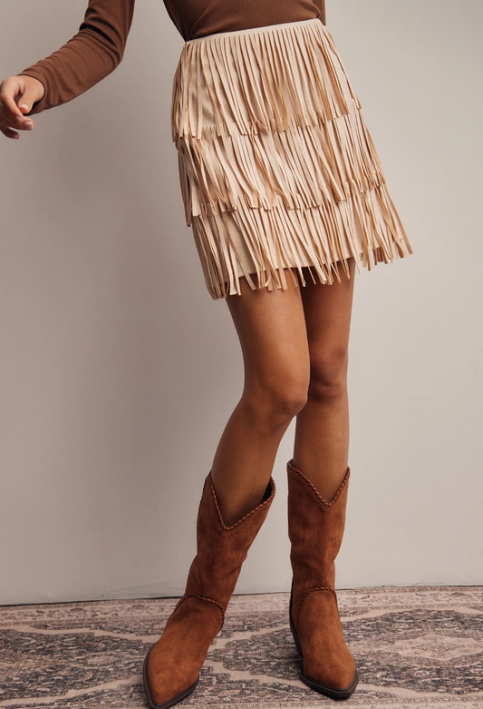 woman wearing a brown top and champagne fringe skirt with brown boots on a neutral background