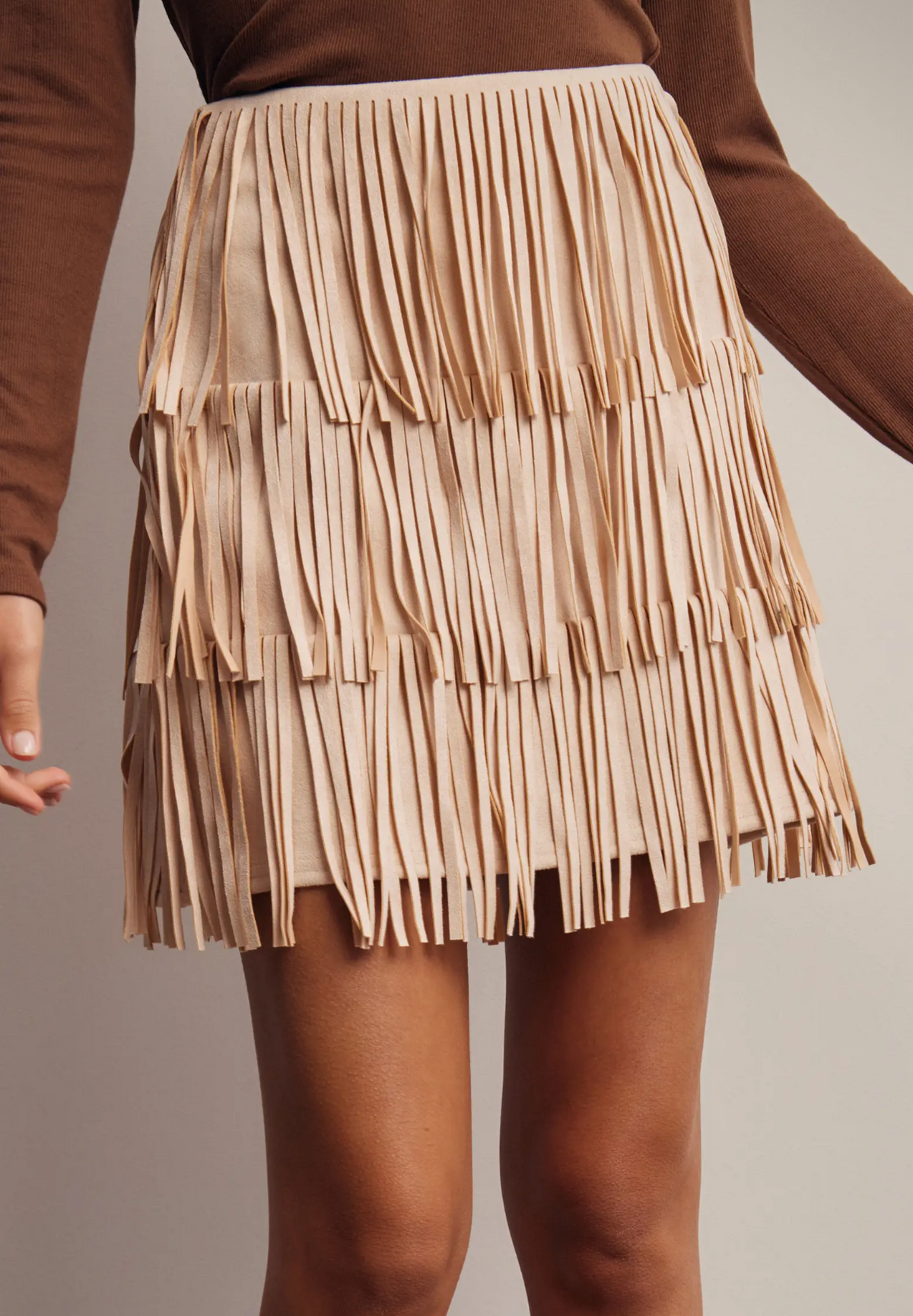 woman wearing a brown top and champagne fringe skirt with brown boots on a neutral background