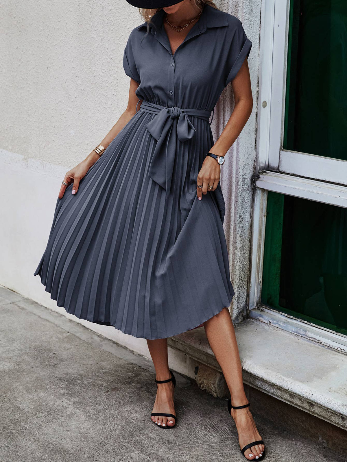 A woman wearing a dark slate gray midi dress with a collar, short sleeves, and a pleated skirt with a tie waist, standing next to a window.