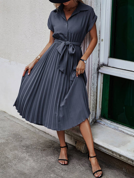 A woman wearing a dark slate gray midi dress with a collar, short sleeves, and a pleated skirt with a tie waist, standing next to a window.