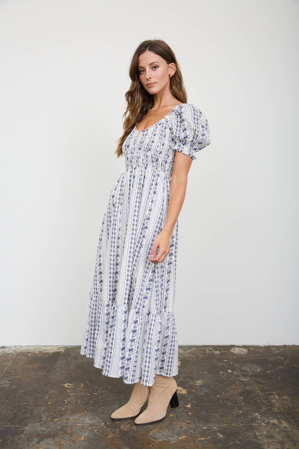 A woman modeling a white midi dress with a black floral print, featuring puff sleeves and a tiered skirt, paired with beige ankle boots against a plain studio background.