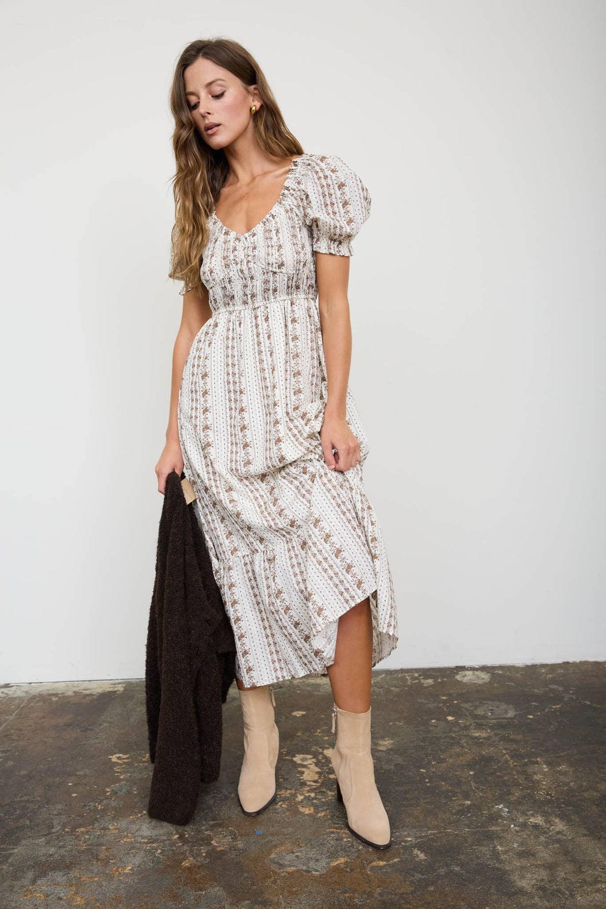 A woman models a white puff-sleeve midi dress with a delicate brown floral print, holding a dark brown cardigan. She is wearing beige ankle boots and standing against a white wall with a rustic concrete floor.