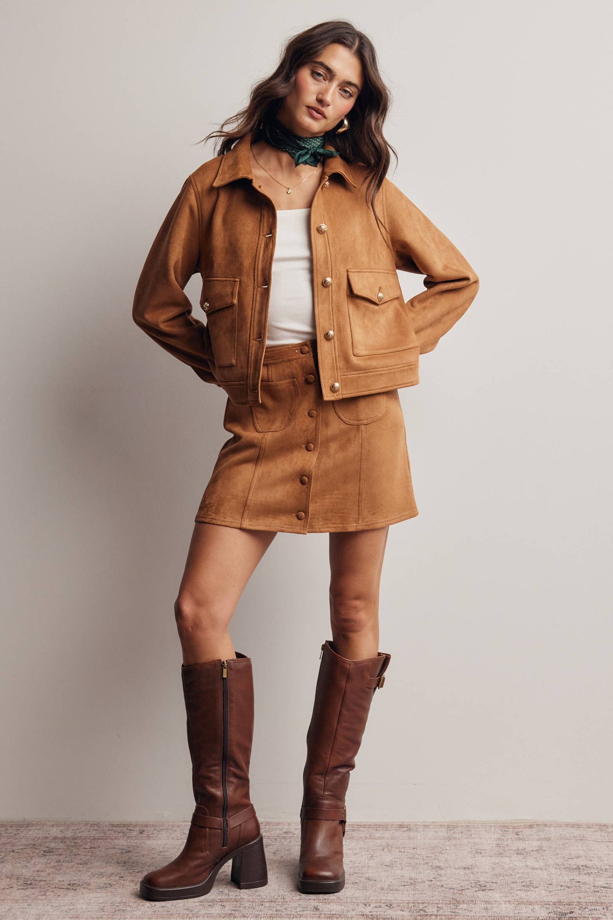Woman wearing a brown suede jacket, skirt, and knee-high boots against a plain background