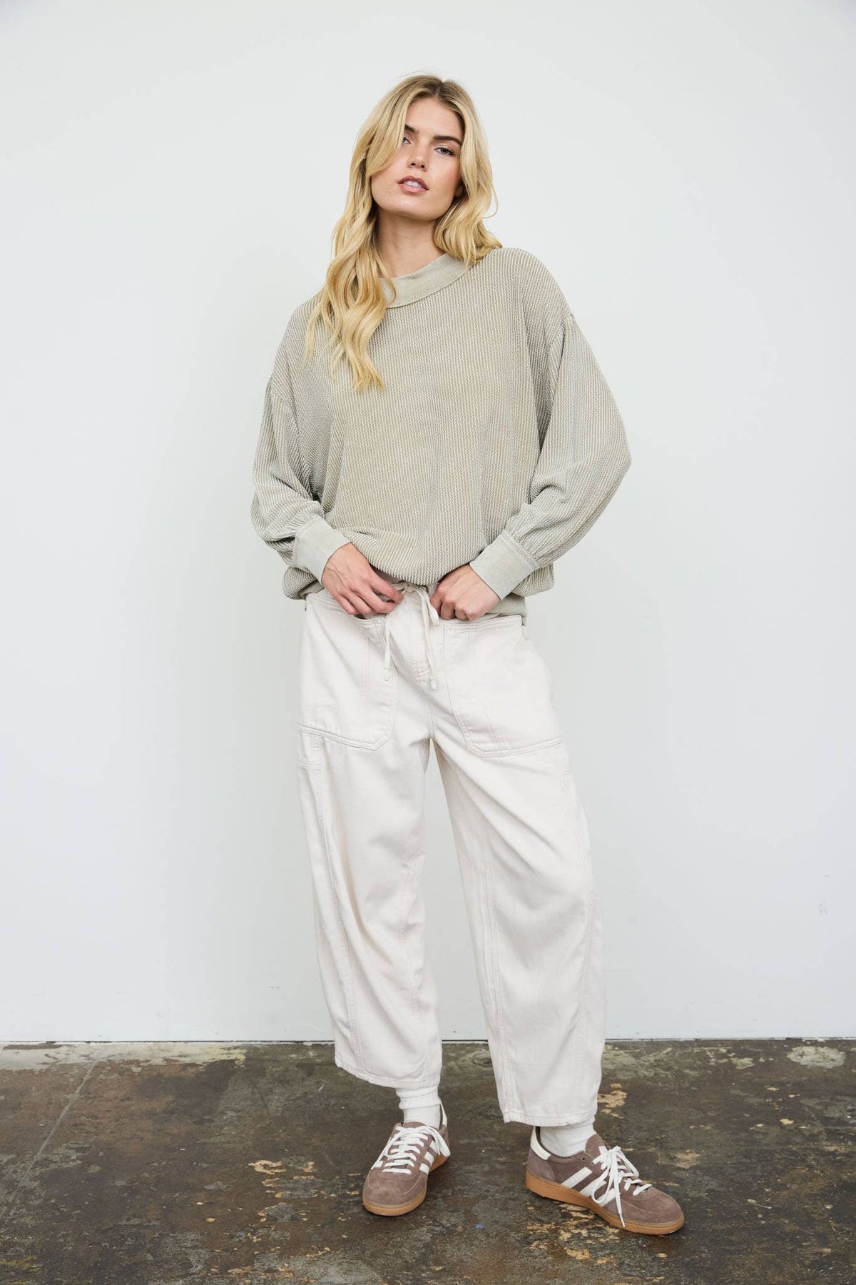 A woman modeling a light beige oversized long-sleeve sweatshirt and cream wide-leg cargo pants, paired with brown sneakers, standing against a white wall.