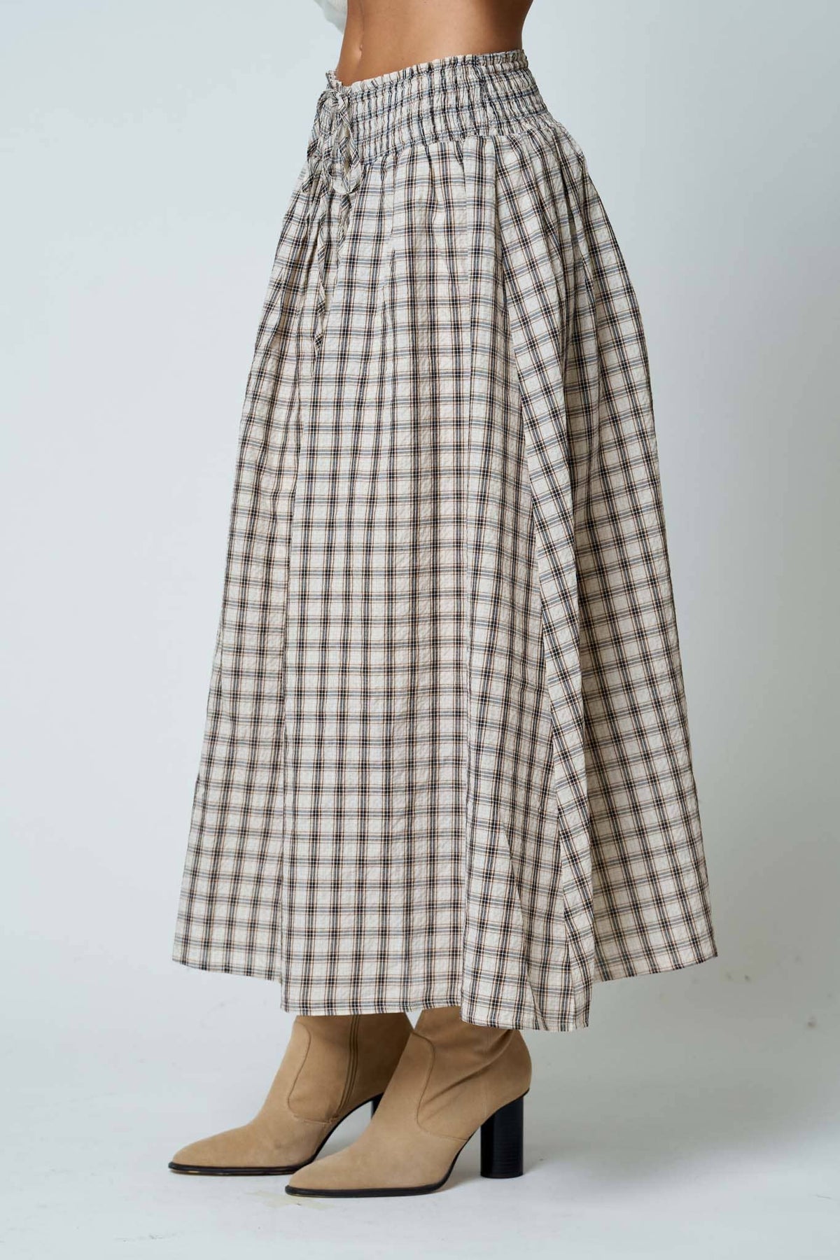 A model is shown from the waist down wearing a high-waisted, pleated A-line midi skirt in a brown and beige gingham plaid pattern, paired with beige ankle boots against a light gray studio background.