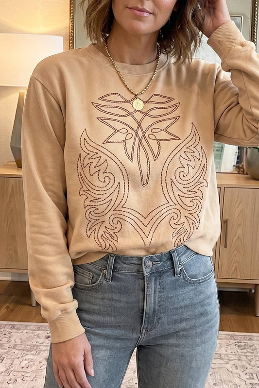 A woman wearing a beige crewneck sweatshirt with a brown embroidered floral and scroll pattern on the front, paired with blue jeans in an indoor setting.