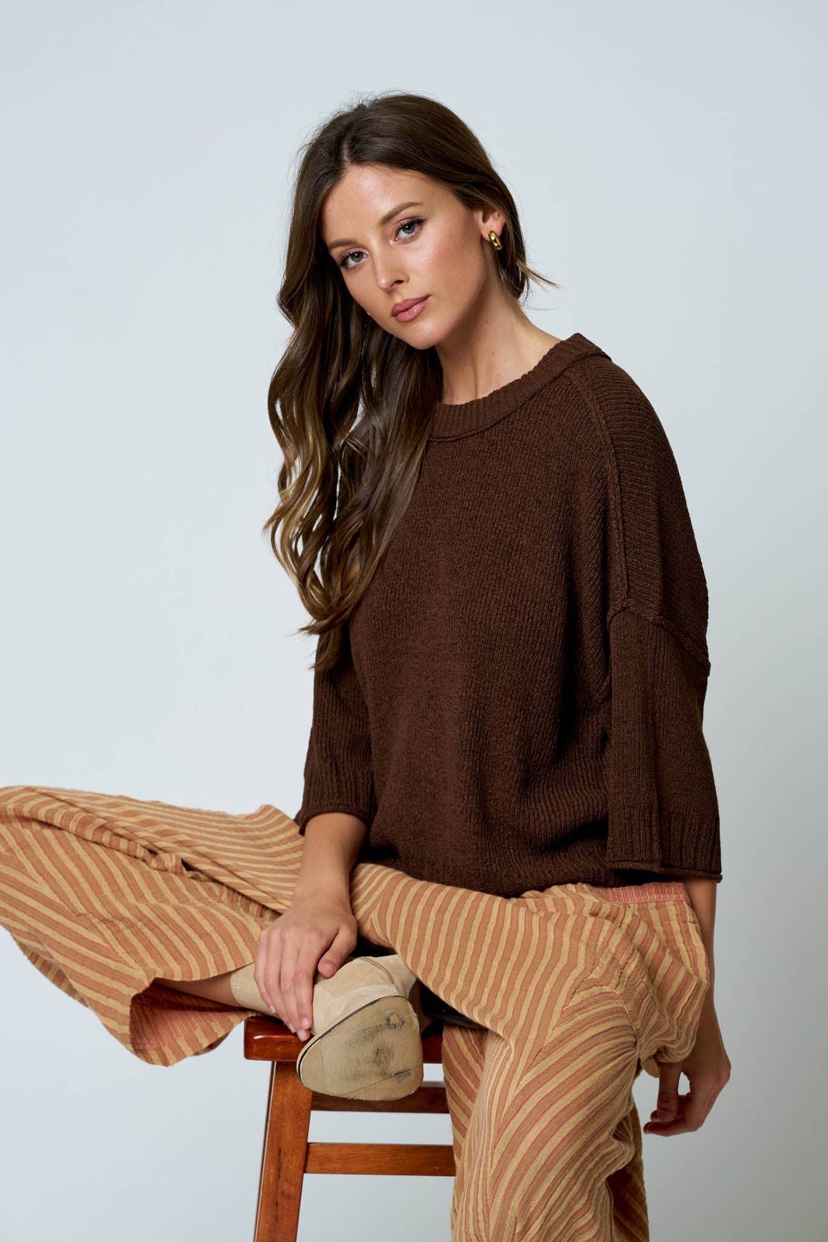 Woman wearing a brown sweater and beige pants sitting on a wooden stool against a light gray background