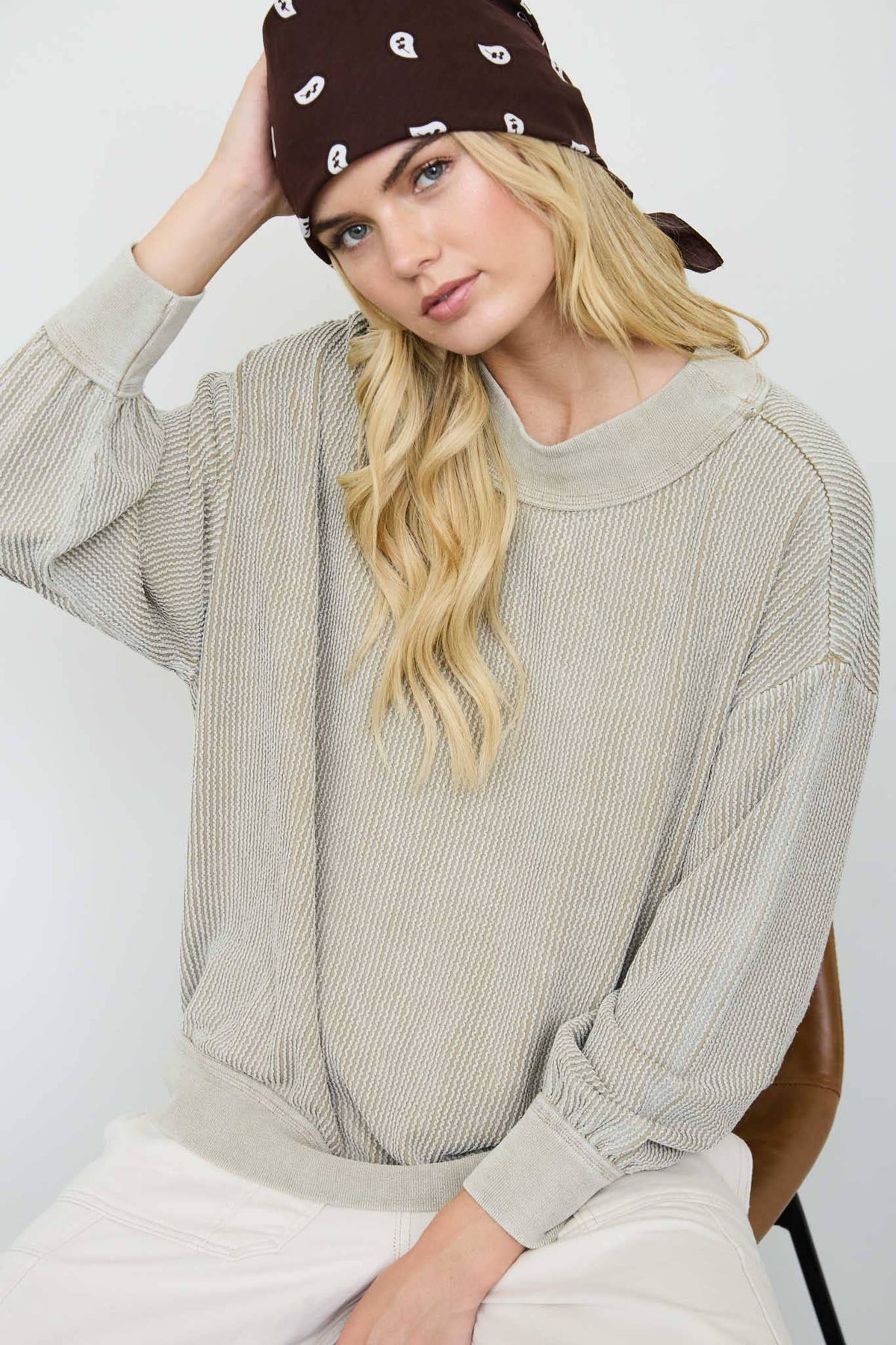 A woman wearing a beige ribbed knit long-sleeve sweater, white pants, and a brown paisley bandana, seated against a light gray background.