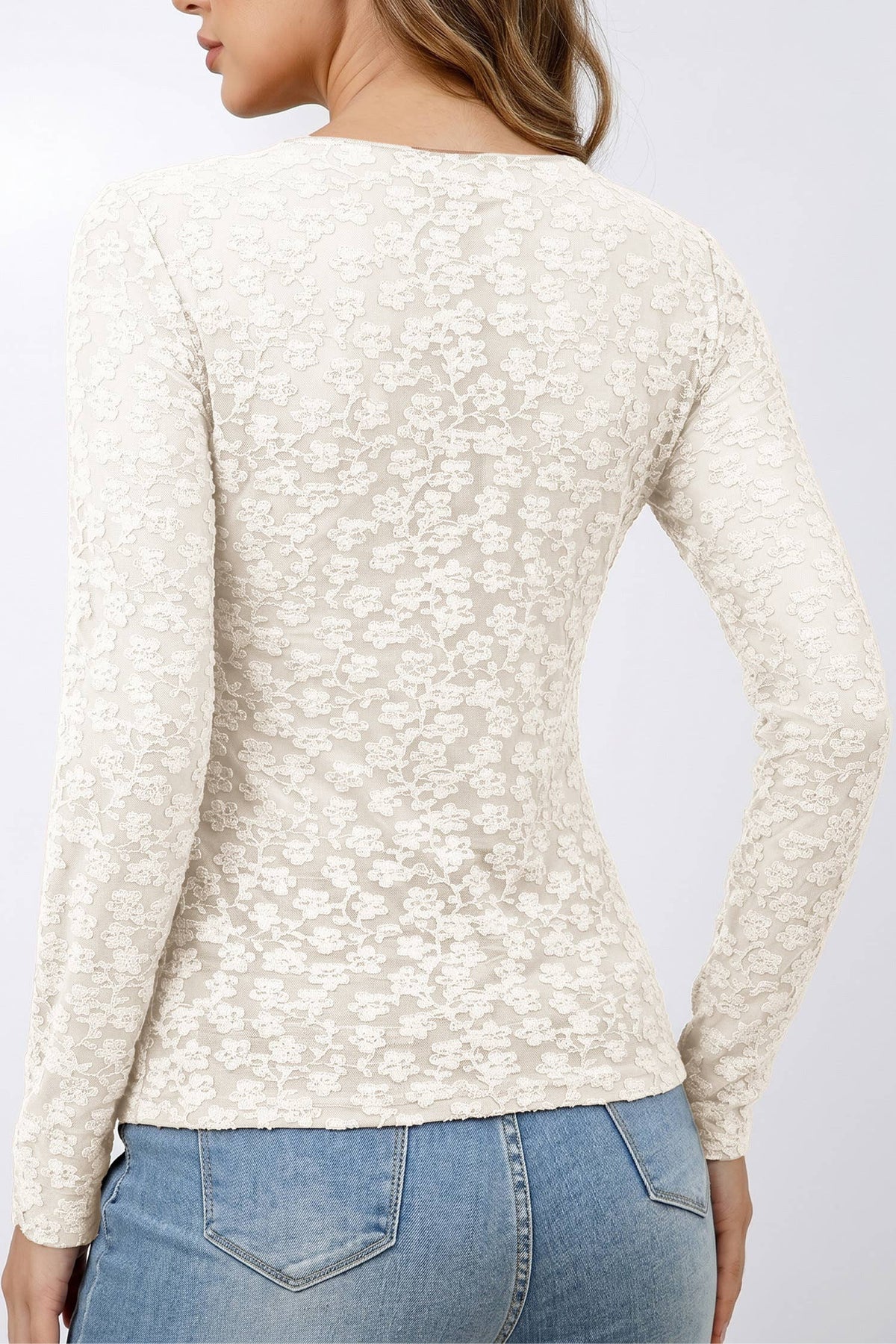 A woman shown from the back wearing a fitted long-sleeve cream lace top and blue jeans against a plain light gray studio background.