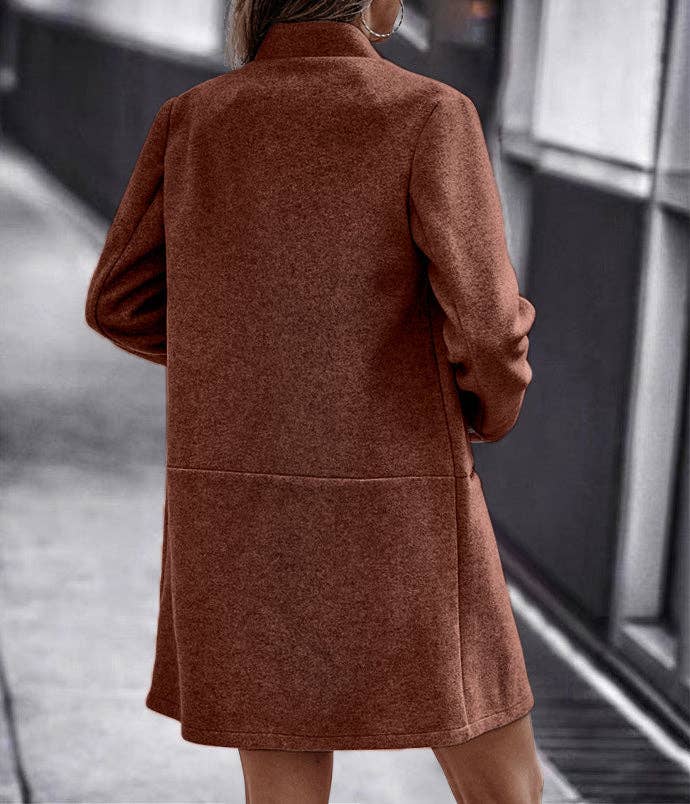 A woman viewed from behind wearing a mid-length rust-brown coat with long sleeves and a high collar, standing outdoors near a modern building.