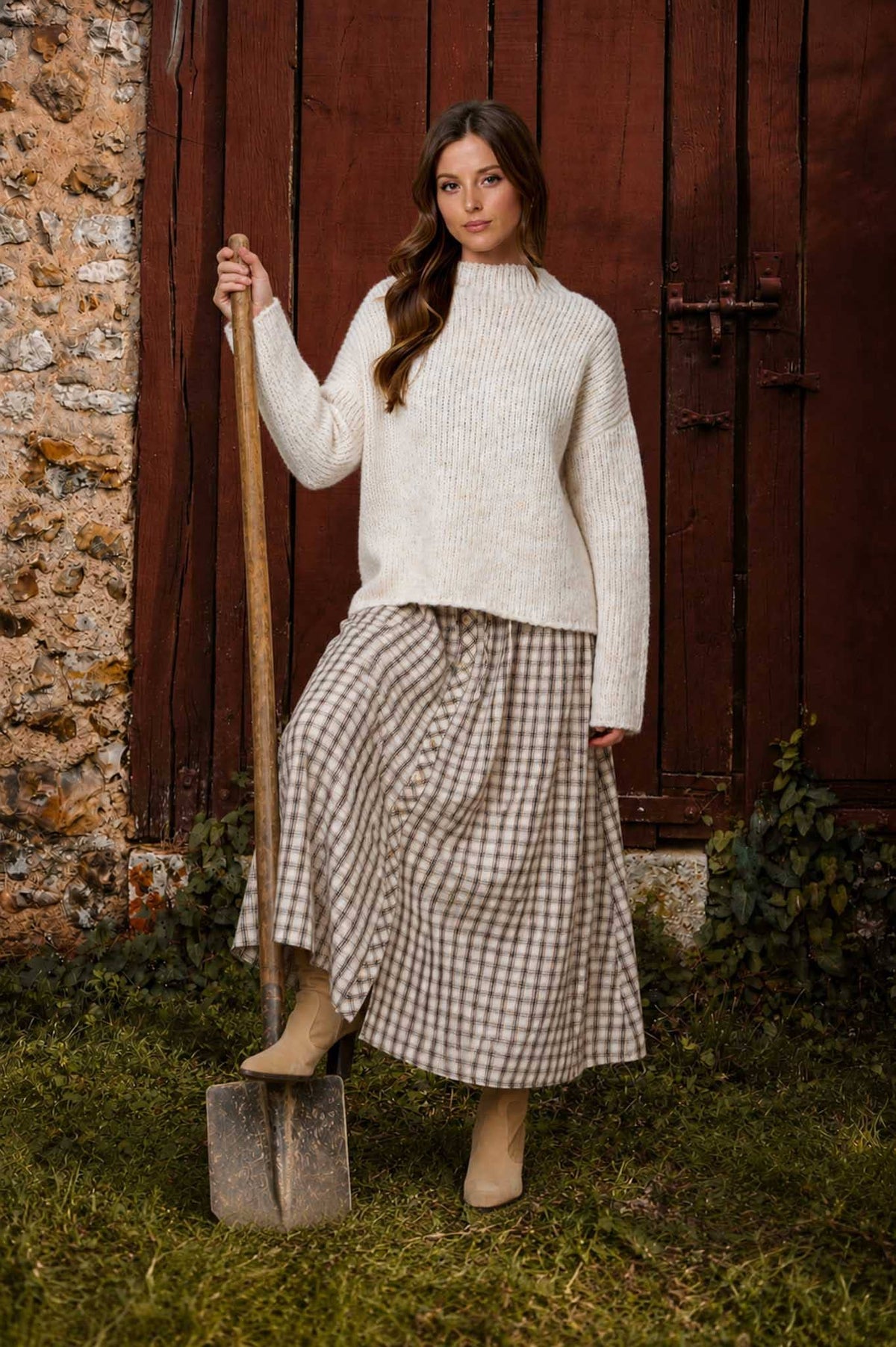 Woman in a white sweater and plaid skirt holding a shovel against a rustic background
