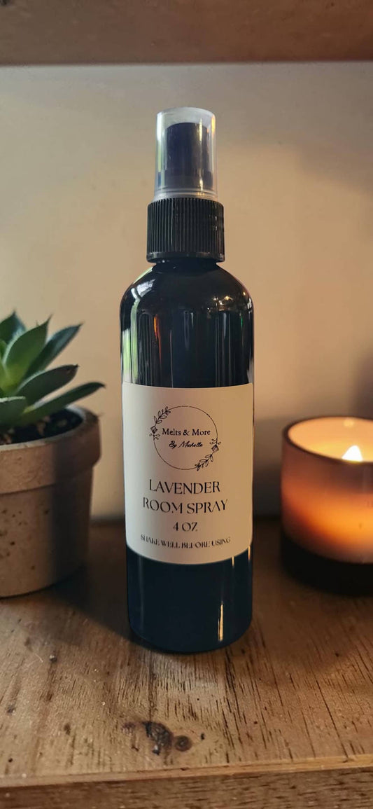 A dark amber glass spray bottle of Lavender roomspray with a white label, set on a wooden shelf next to a lit candle and a small succulent plant.