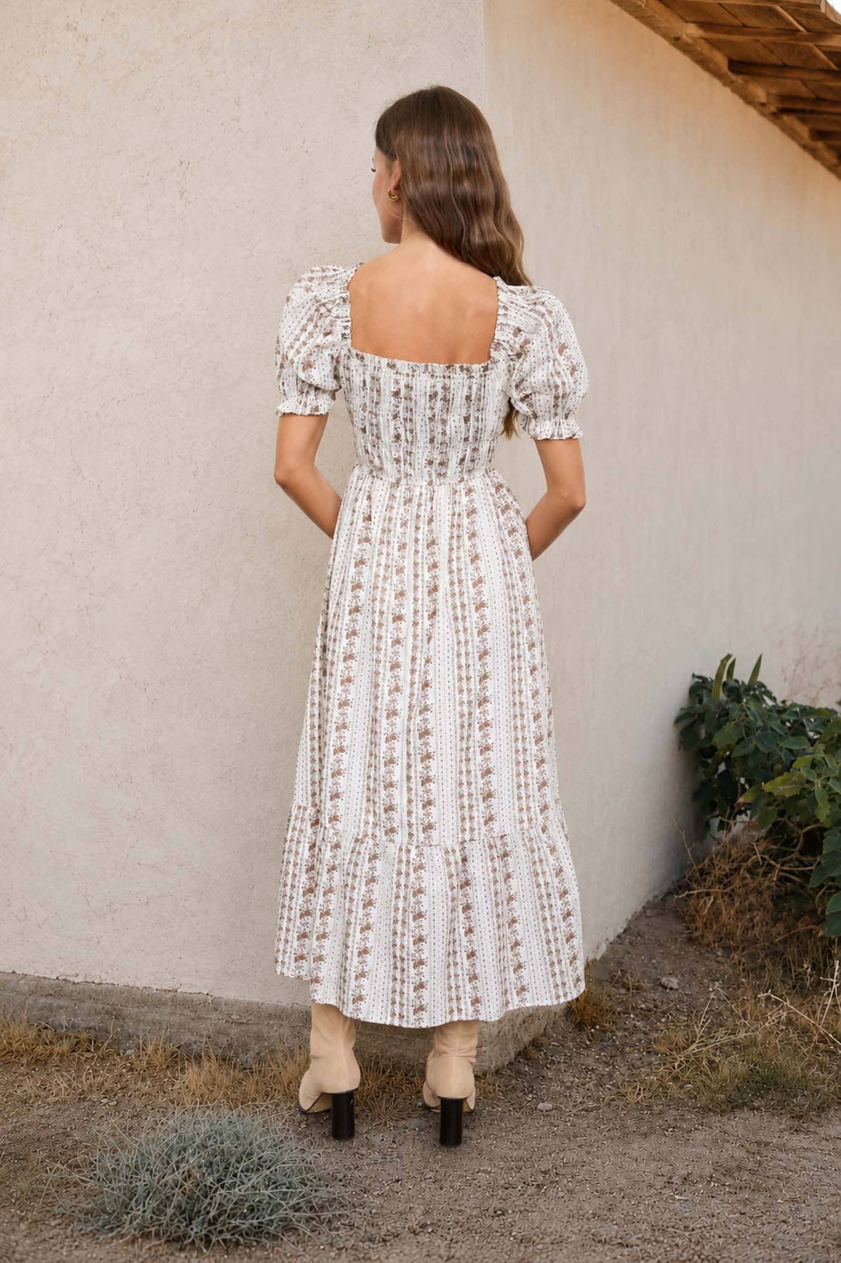 Woman in a white patterned dress standing against a beige wall with plants around.