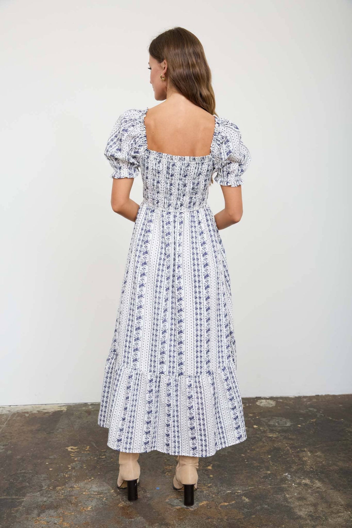 A woman viewed from the back wearing a white midi dress with a blue vertical floral pattern, puff sleeves, and an open back, standing against a plain light wall.