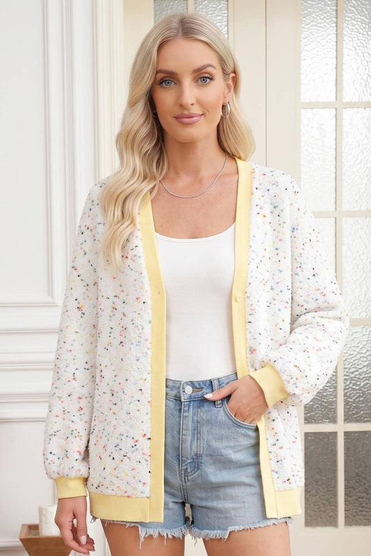 A woman wearing a white open-front cardigan with a multicolor speckle pattern and yellow trim, styled over a white tank top and denim shorts.
