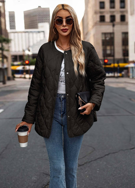 A woman walking on a city street wearing a dark olive green quilted bomber jacket over a white graphic tee and blue jeans, holding a coffee cup and a black clutch.