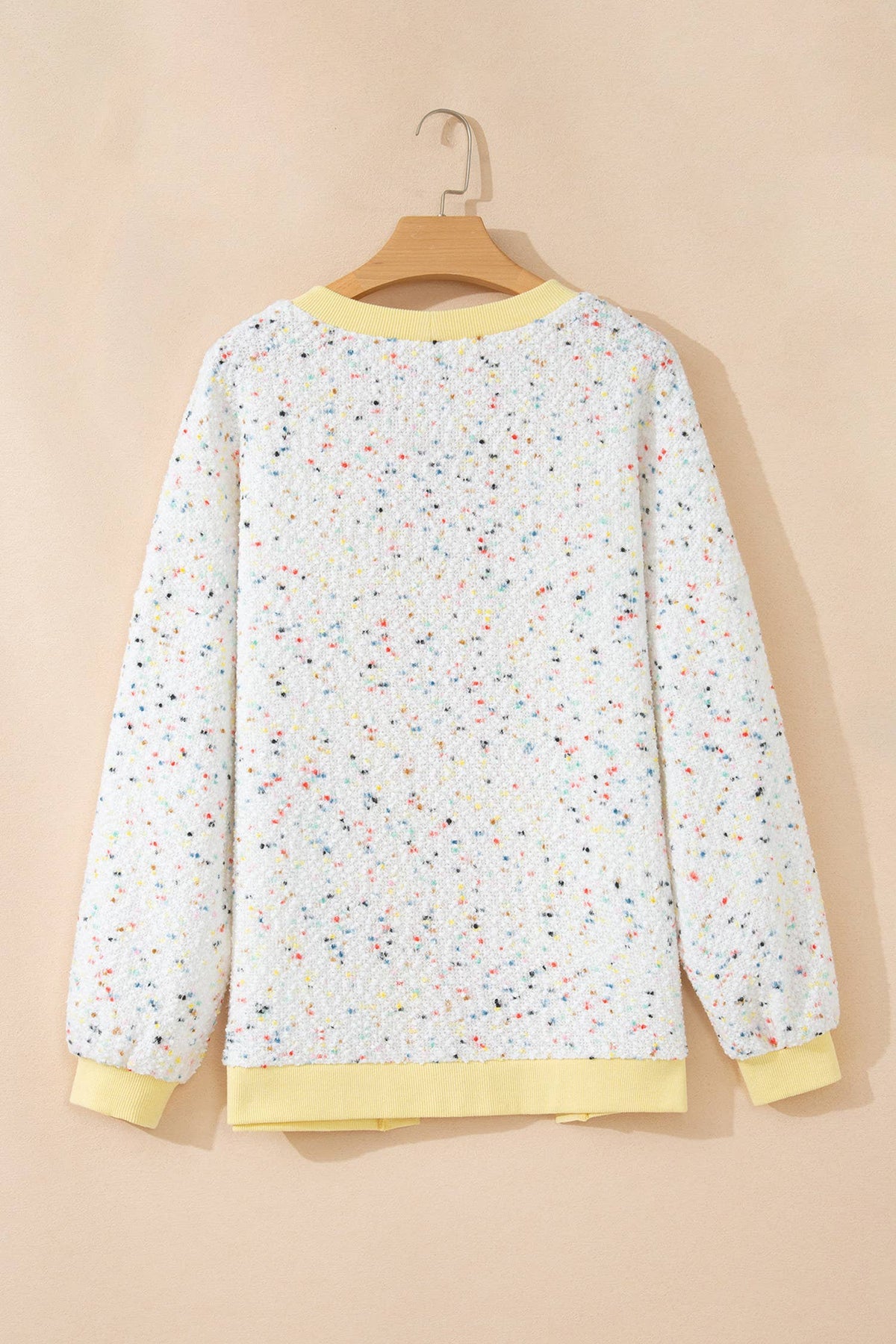 A long-sleeve crewneck sweatshirt with a white base covered in a multicolor speckle pattern, featuring solid pale yellow ribbed trim at the collar, cuffs, and hem, hanging on a wooden hanger against a solid beige background.