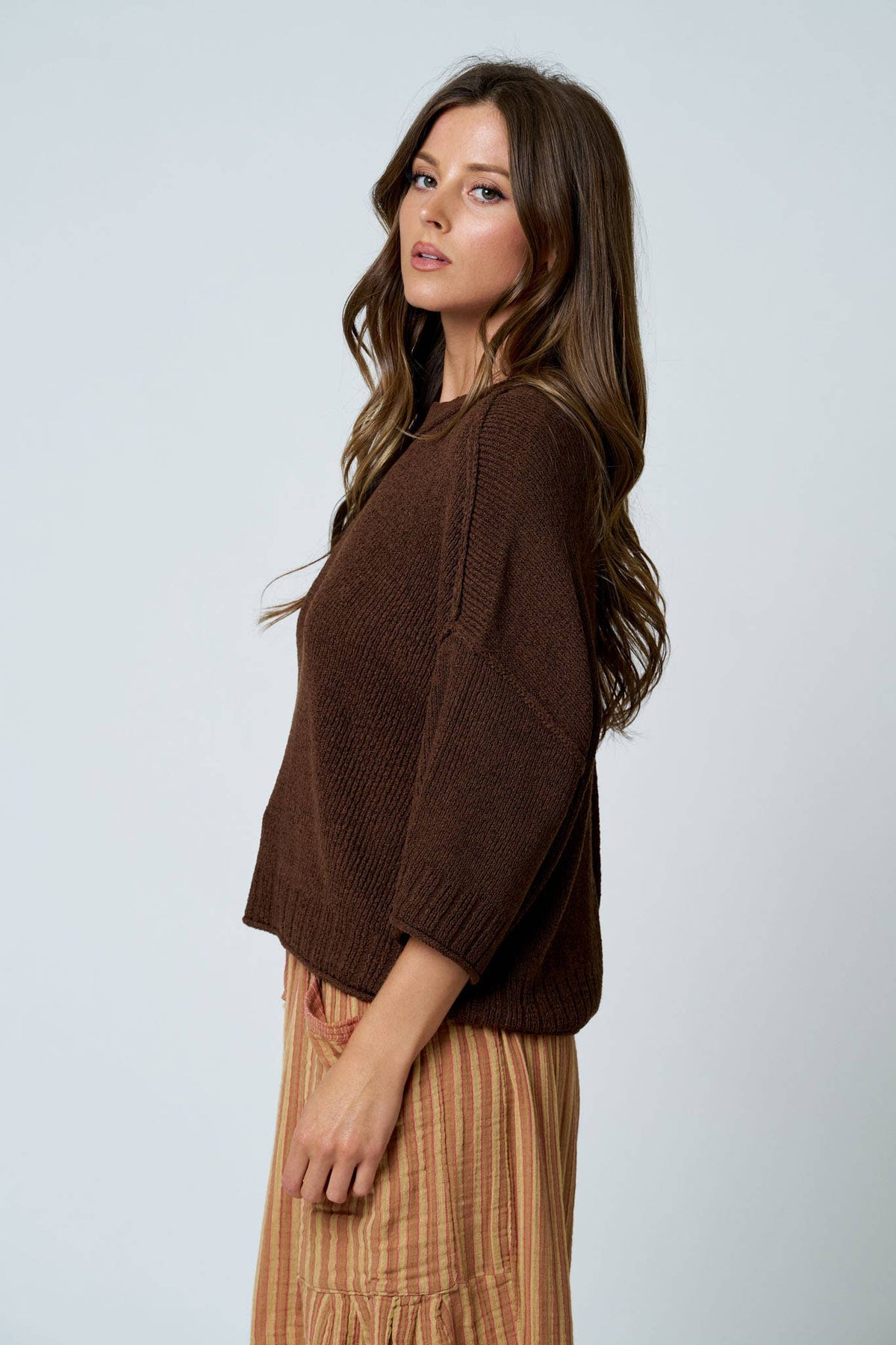 Woman wearing a brown sweater and patterned pants against a light gray background