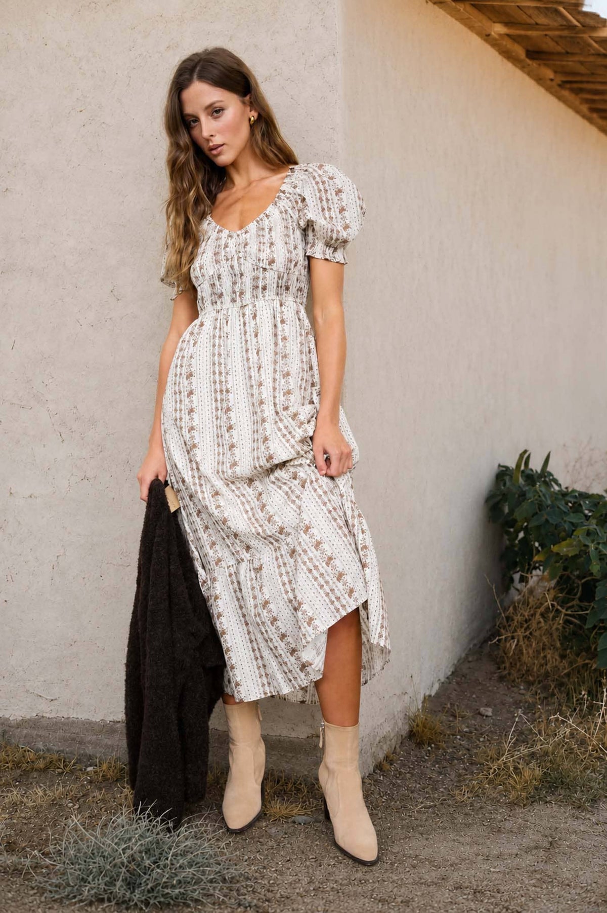 Woman in a patterned dress standing against a plain wall with plants around.