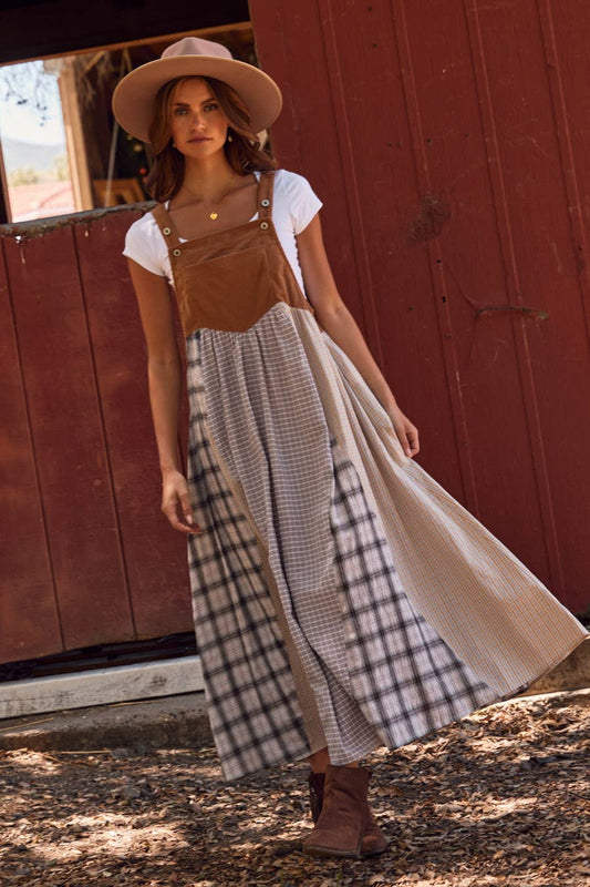 Woman wearing a brown overall dress over a white shirt and plaid skirt, standing in front of a red wooden door.