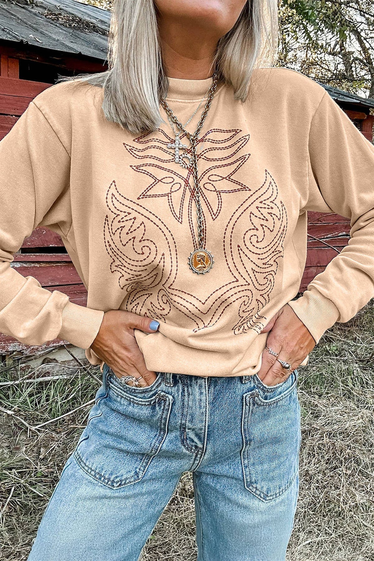 A woman wearing a beige crewneck sweatshirt with a brown embroidered western-style wing design on the front, paired with blue jeans, standing outdoors in front of a rustic red wooden structure.