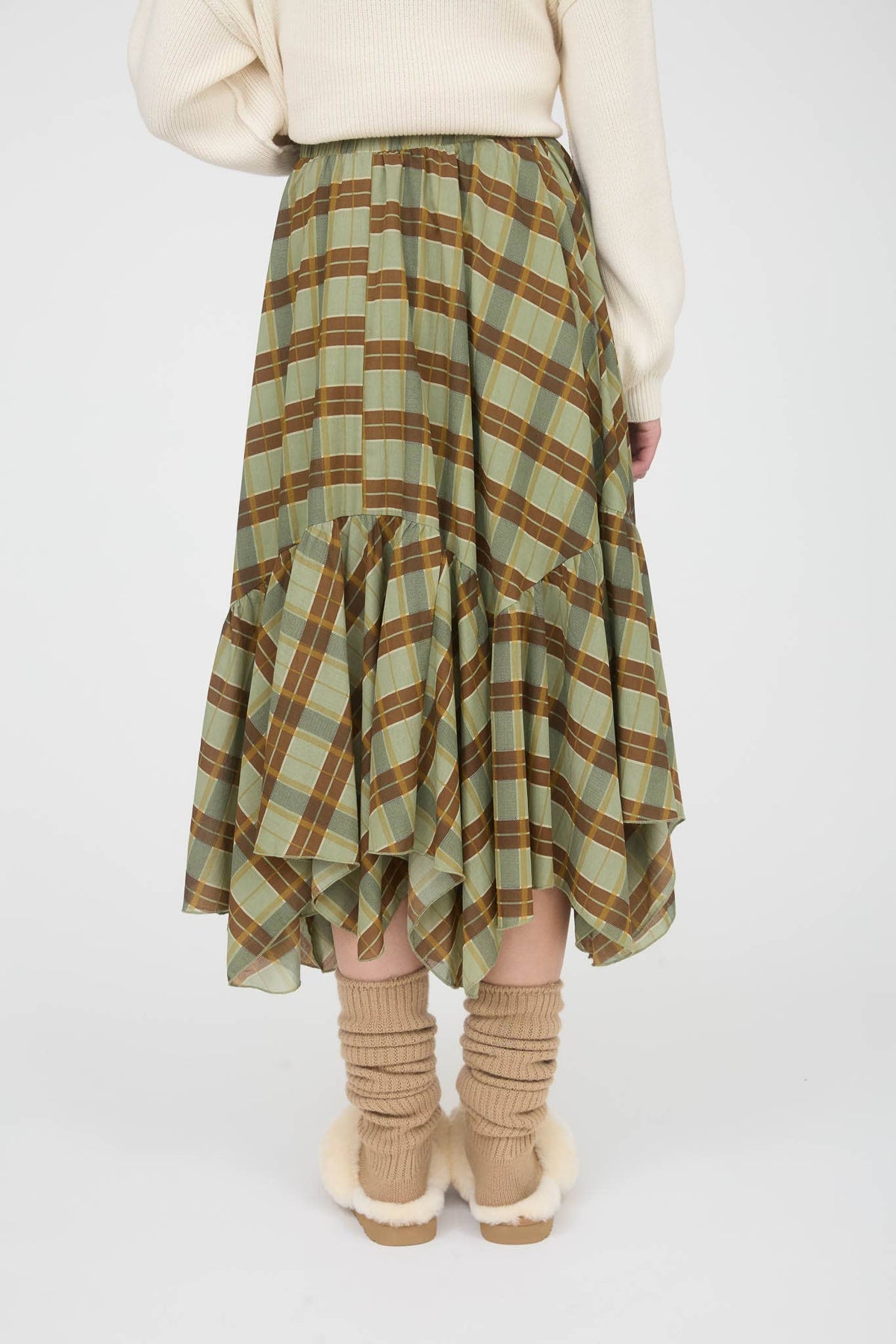 A person shown from the back wearing a green and brown plaid tiered midi skirt with an asymmetrical hem, styled with a cream sweater, beige knit socks, and beige slippers on a light gray studio background.