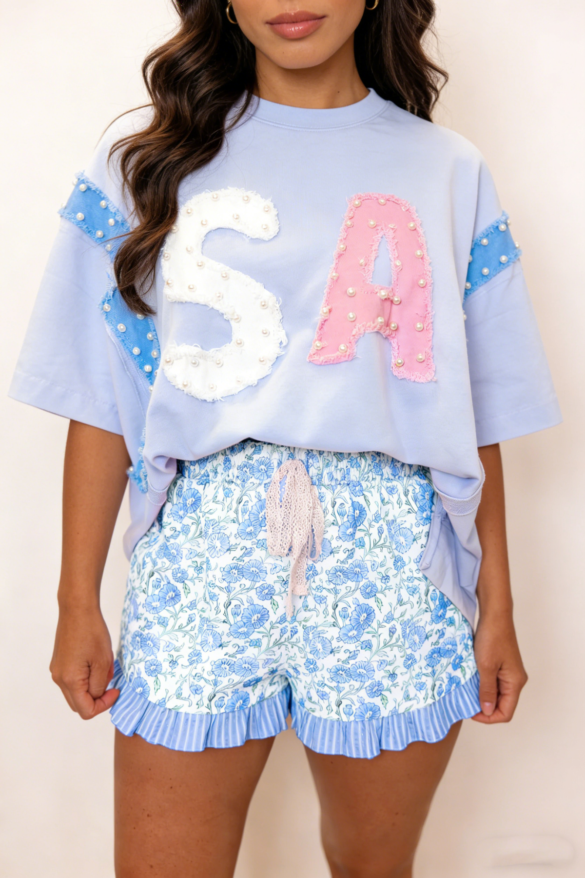 Woman wearing a light blue oversized shirt with 'USA' letters and floral shorts on a plain background