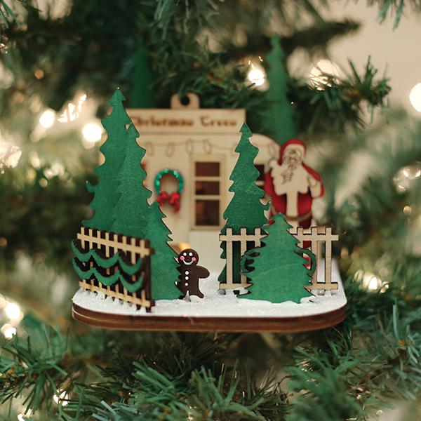 Chrismas tree lot ornamnet with  christmas tree, gingerbread man, Santa, and fence wooden figures hanging from a blurred tree branch.